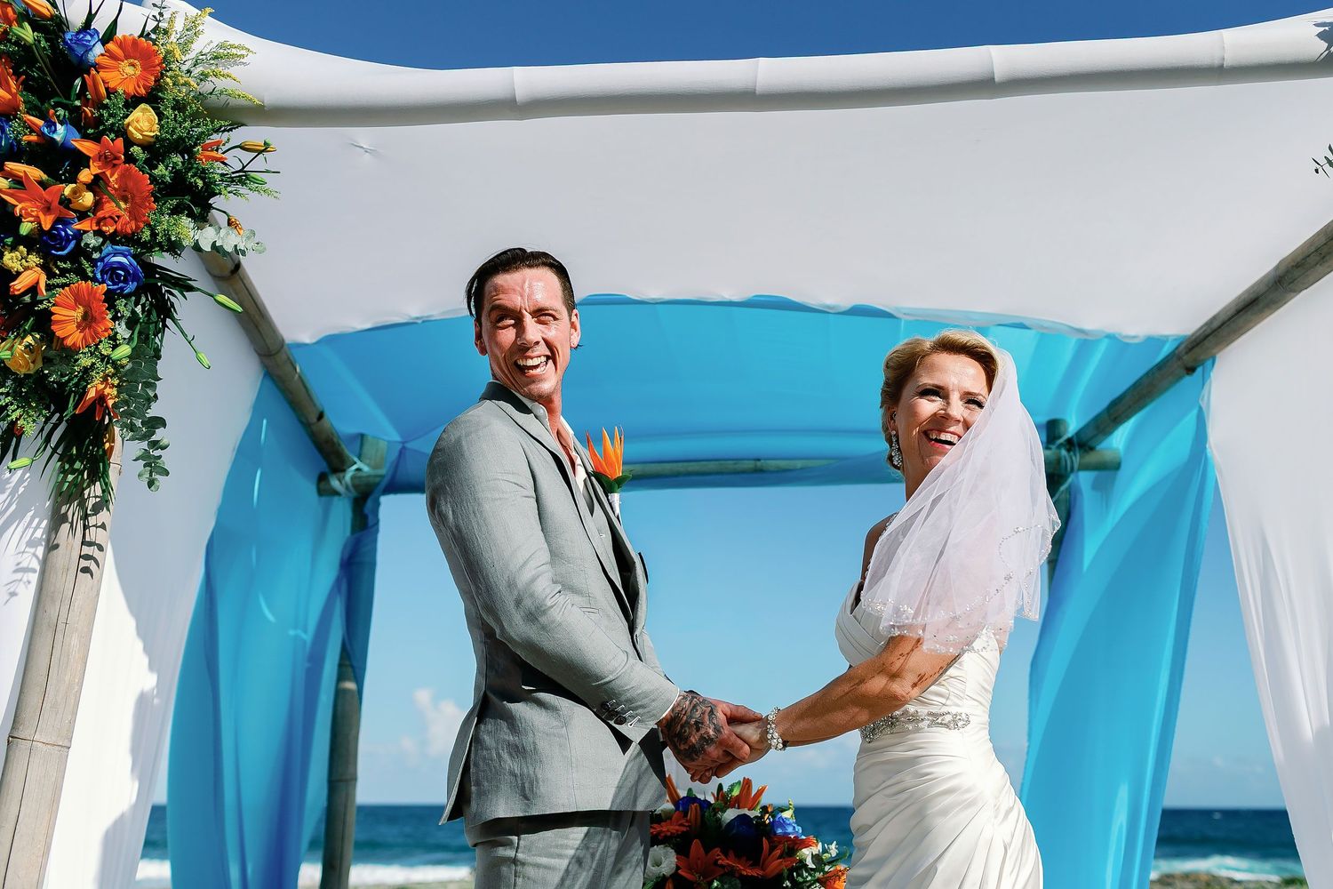 Carly and Scott holding hands beneath their colorful beach wedding canopy