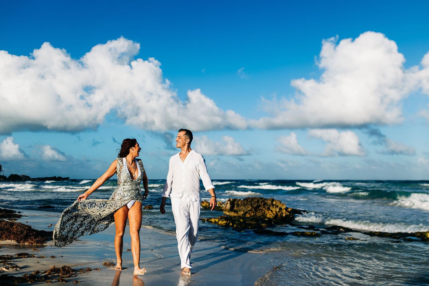 Sarah and Evish walk barefoot beside the Caribbean Sea beneath dramatic clouds.