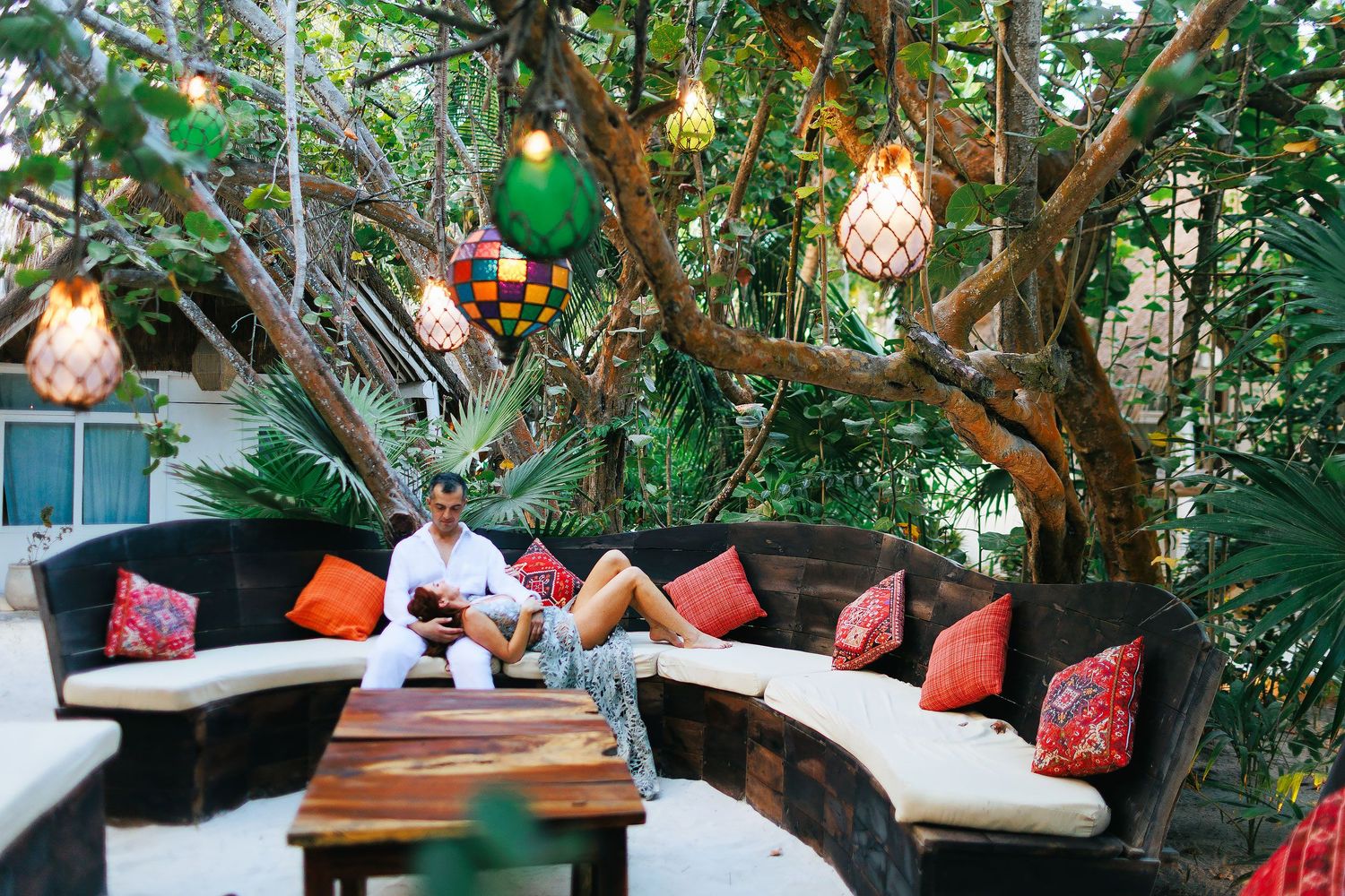 Sarah rests across Evish’s lap beneath colorful lanterns in a tropical lounge.
