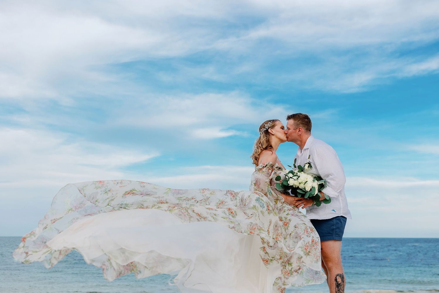 Autumn’s floral wedding dress flowing in the ocean breeze as she kisses Brian on the Riviera Maya beach.