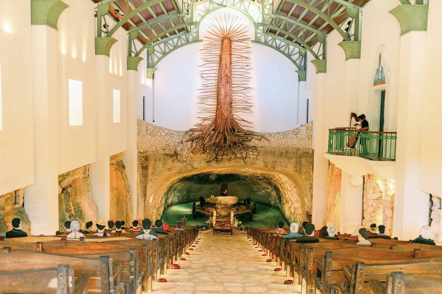 A wide indoor view of the Capilla de Guadalupe at Xcaret Park, featuring a stone altar floating over a cenote and a 39-foot tall wood-carved