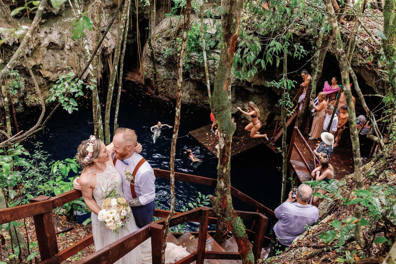 Jeff and Veronica posing on the wooden stairs while wedding guests swim and jump into the water at Cenote Hats’uts near Playa del Carmen.