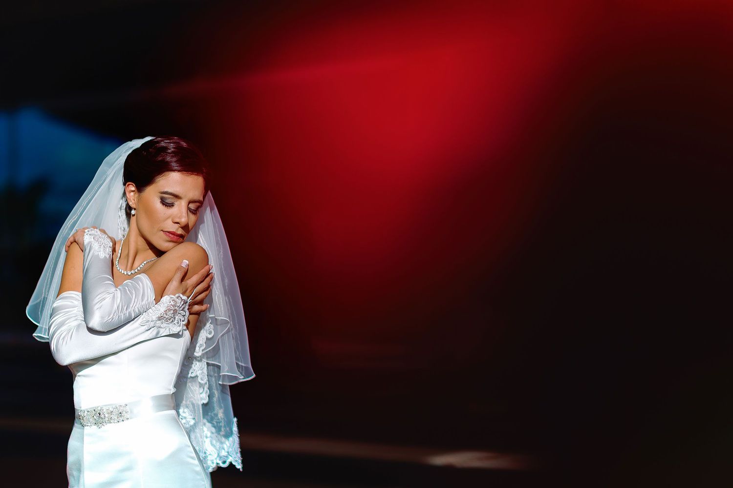 Linda posing with her arms crossed in a white satin wedding dress, long gloves, and lace veil against a dark red background at her Illinois
