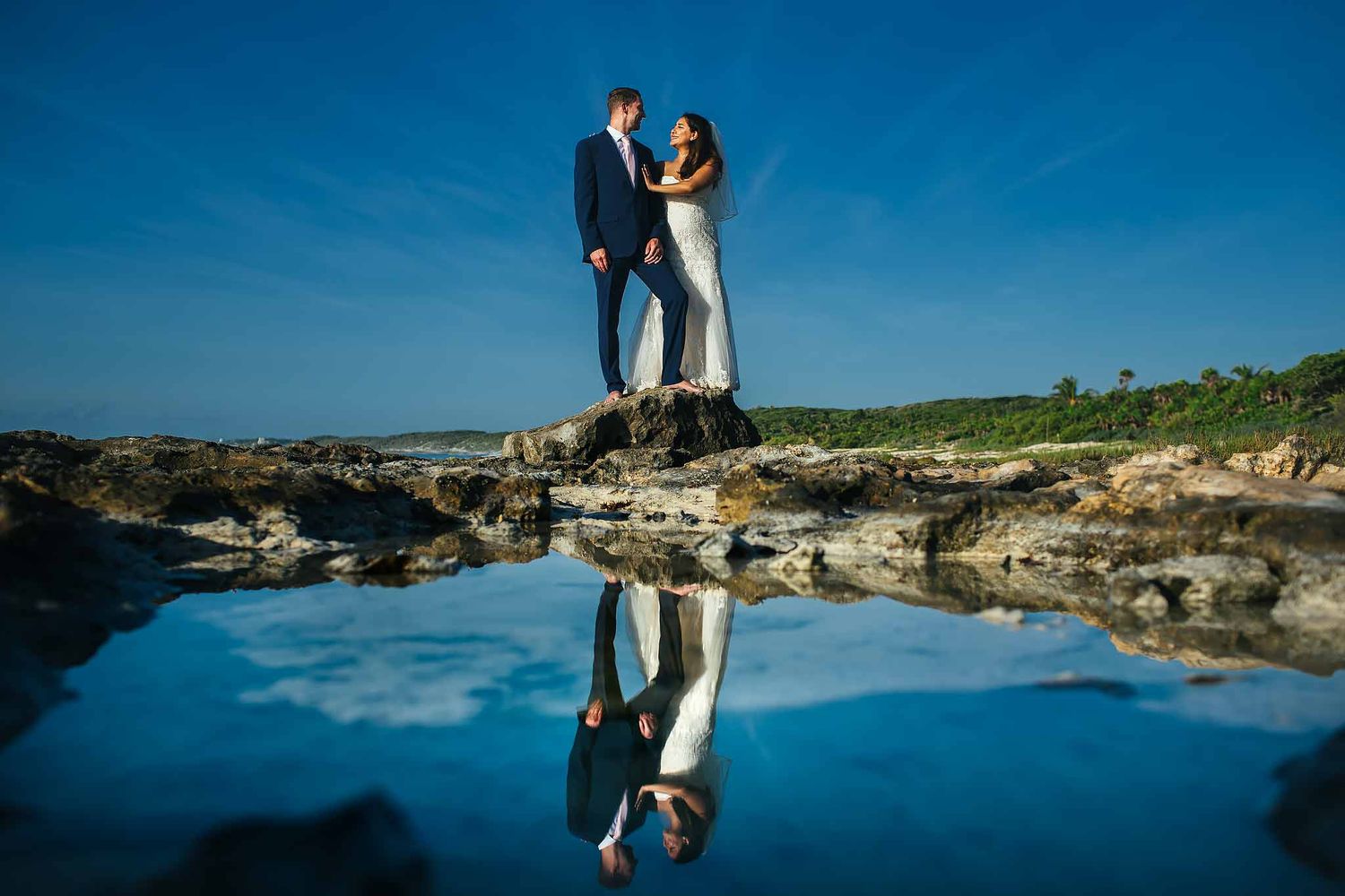 Bride and groom portrait on the rocky shoreline at Secrets Aura Cozumel captured by a Cozumel wedding photographer with ocean reflection.
