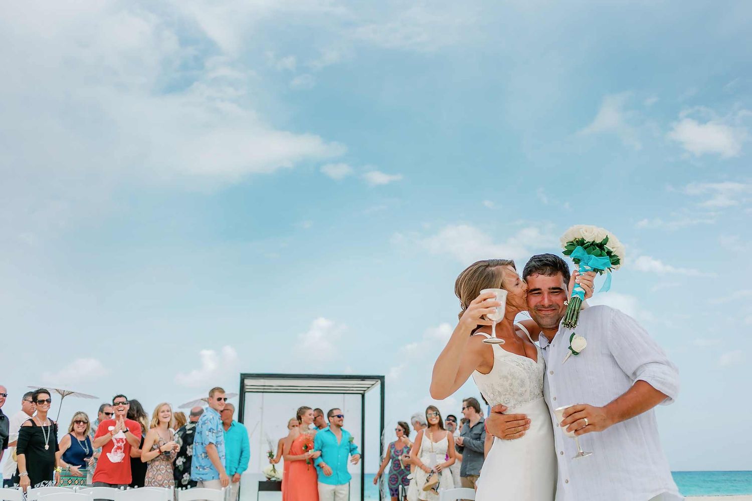 Bride kissing groom during beach wedding celebration in Playa del Carmen with guests and ocean backdrop