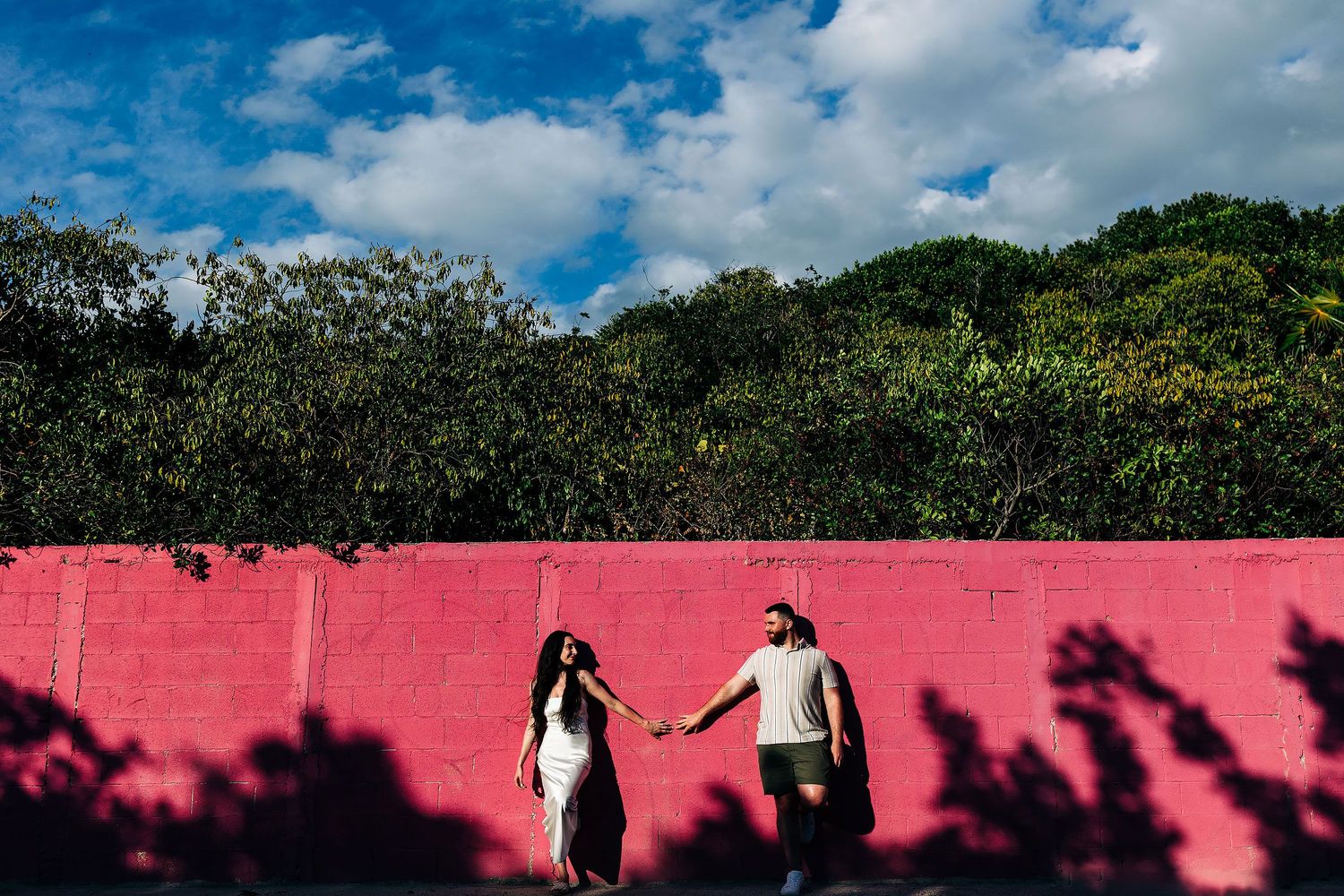 Alexia and Sam hold hands beside a bright pink wall beneath tropical trees in Isla Mujeres.