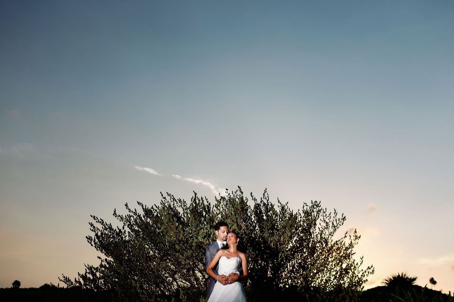 Felicitas and Keith embrace in front of tropical greenery at sunset.