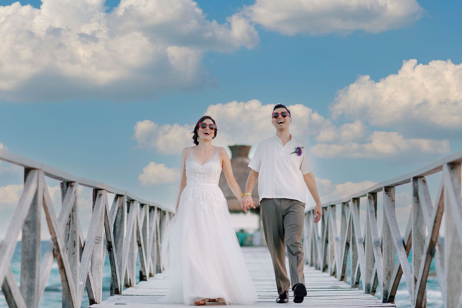 Bride and groom wearing heart-shaped sunglasses while walking hand in hand along a pier during their wedding portraits at Grand Palladium