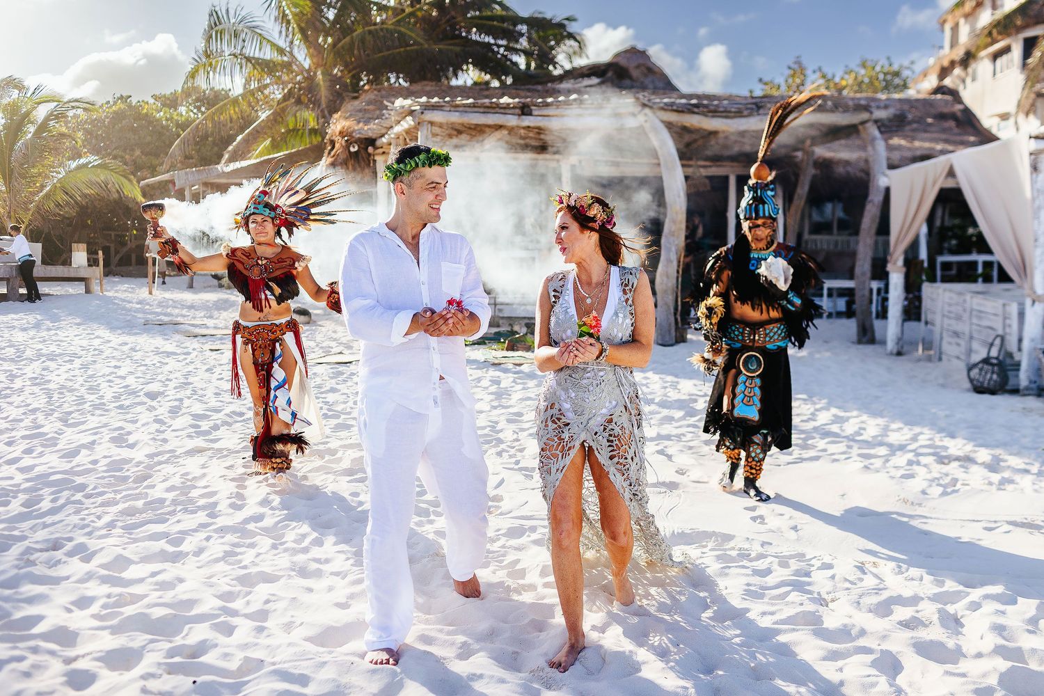 Sarah and Evish walk barefoot on the beach between colorful ceremonial participants.