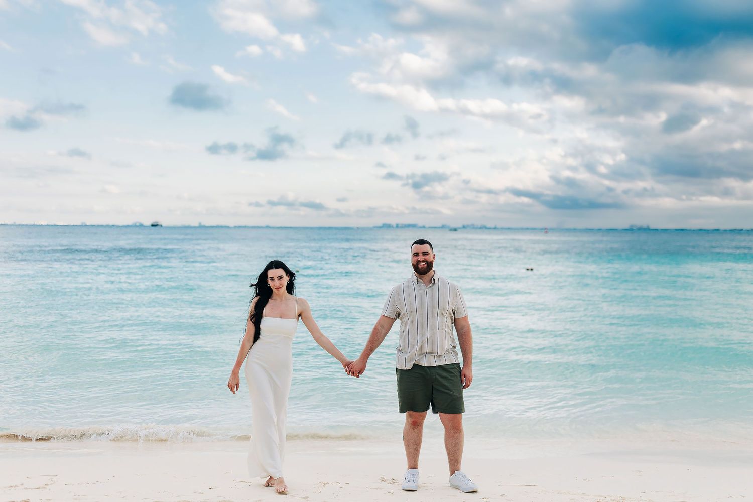 Newly engaged Alexia and Sam hold hands at the edge of a calm turquoise beach in Isla Mujeres.