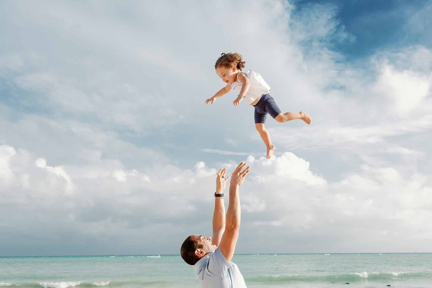 Father lifting child in the air during a playful family photoshoot on a Cancun beach with ocean and blue sky