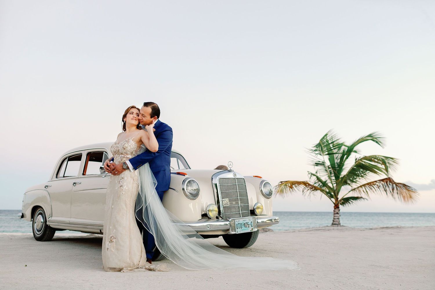 Bride and groom embracing beside a vintage white car on the beach during their wedding portrait session at Blue Venado in Playa del Carmen.