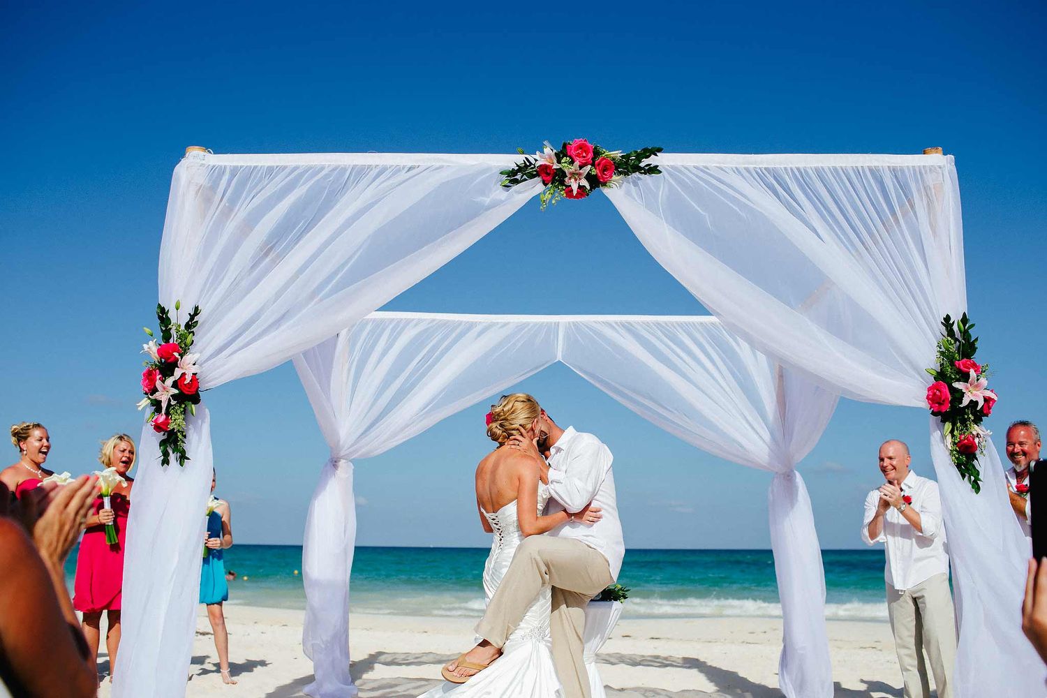 Bride and groom kissing under beach wedding arch in Cancun with guests cheering, destination wedding photography Riviera Maya