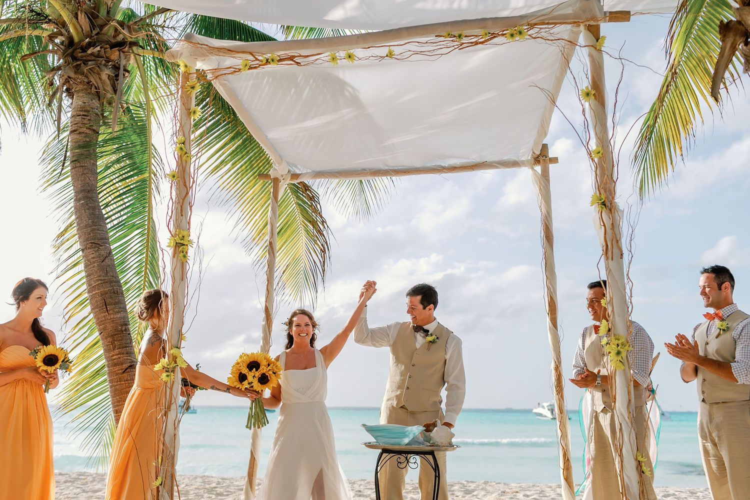 Li and Carlos raise their joined hands as their wedding party celebrates beside the Caribbean.