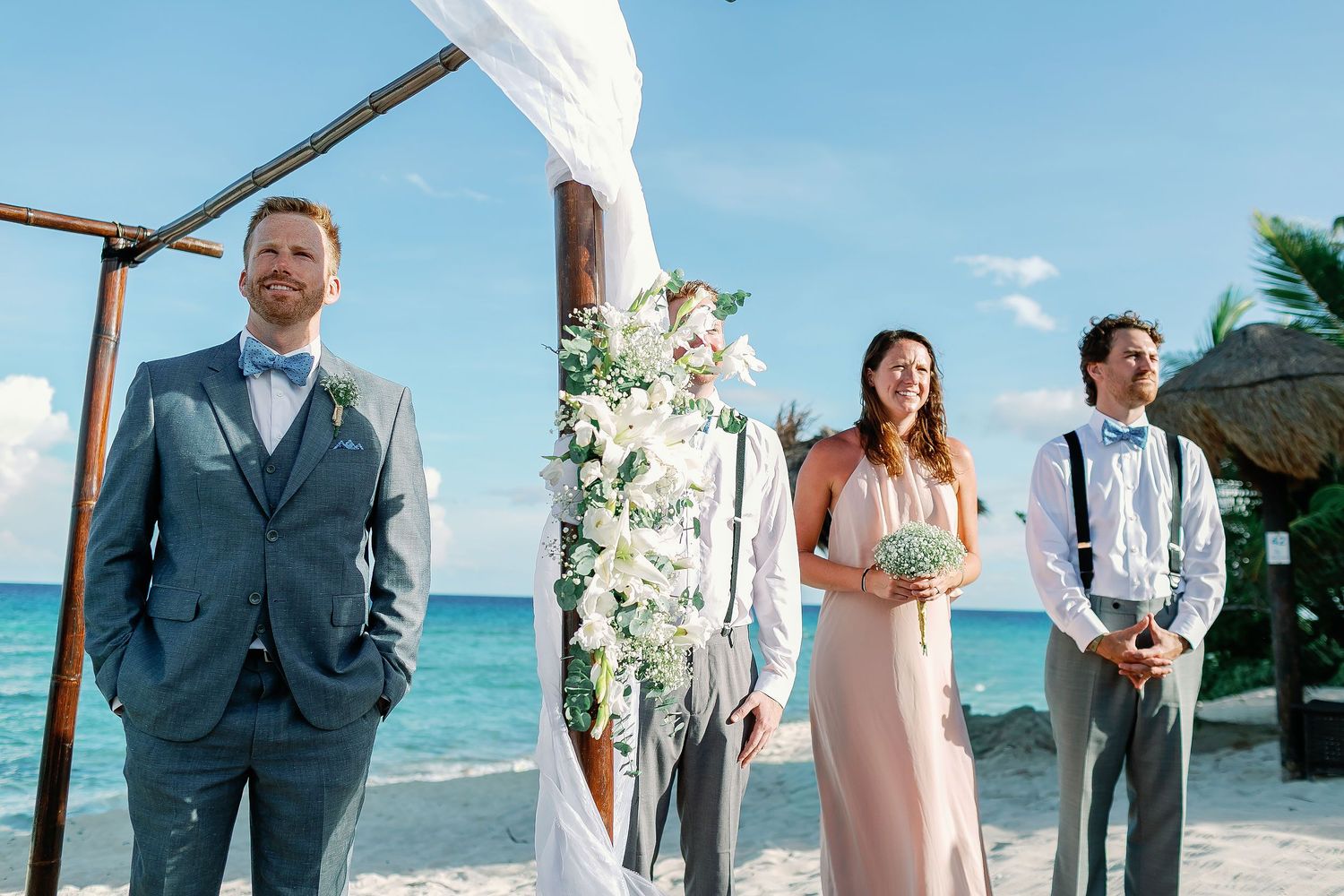 Alan waiting with the wedding party beneath a white floral arch before Cressida arrives at their private Riviera Maya beach ceremony.