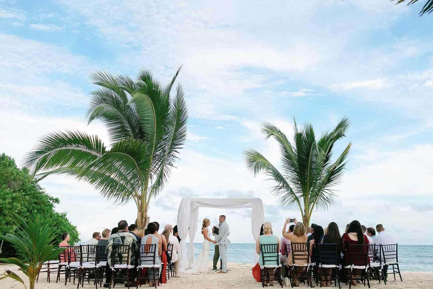 Isla Mujeres beach wedding ceremony with bride and groom under arch, guests seated, palm trees and Caribbean ocean backdrop