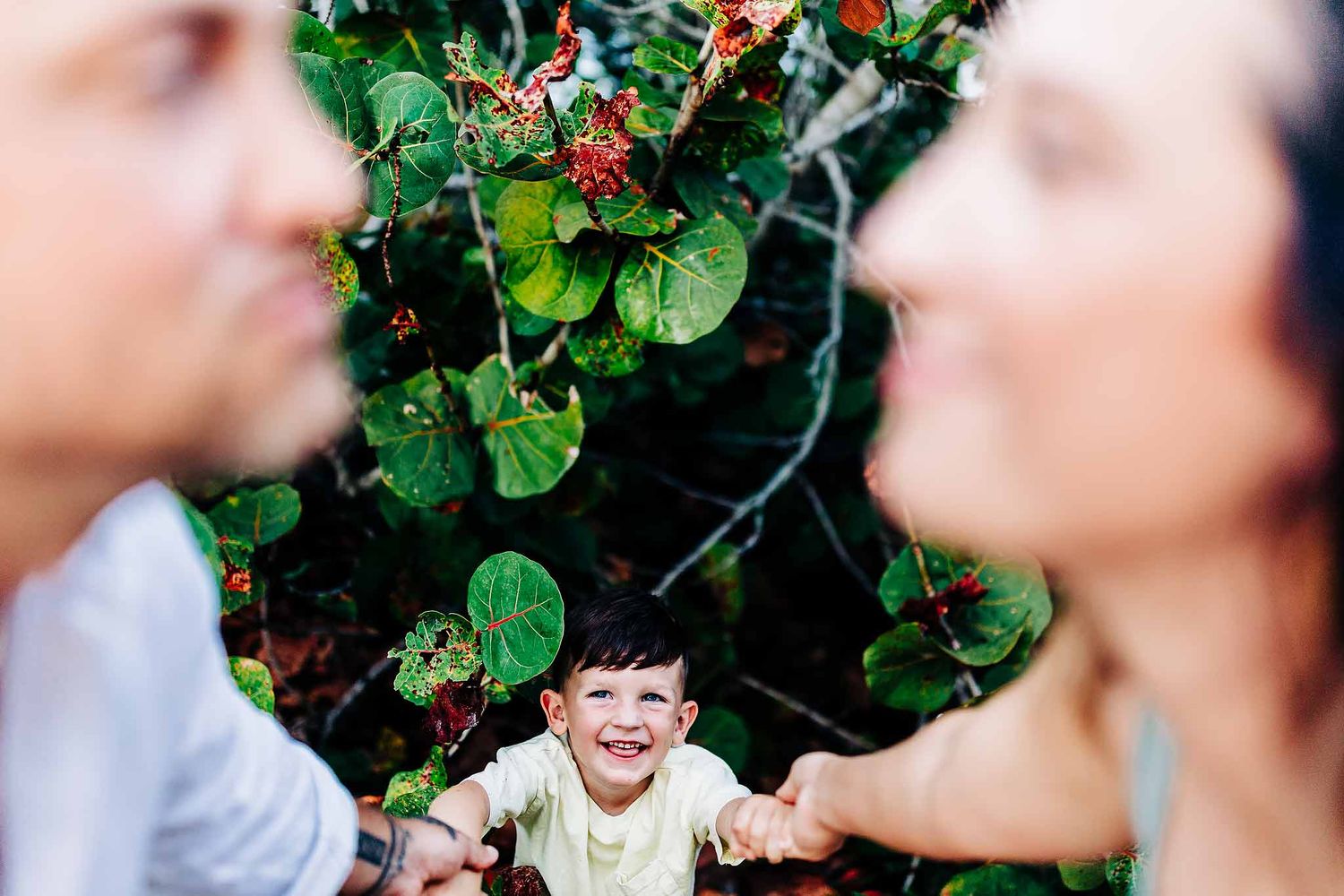 Candid family photoshoot in Cancun with smiling child holding parents hands surrounded by tropical greenery