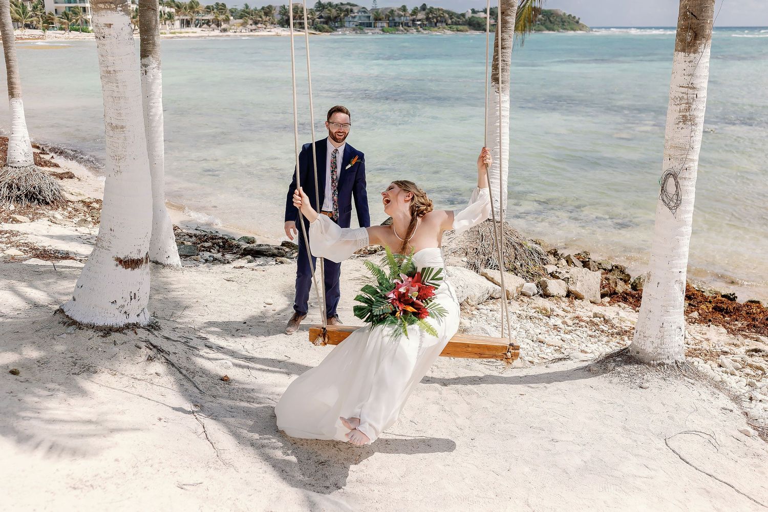 Nicole laughing on a beach swing while Thomas stands behind her during their wedding portraits at Blue Venado in Playa del Carmen.