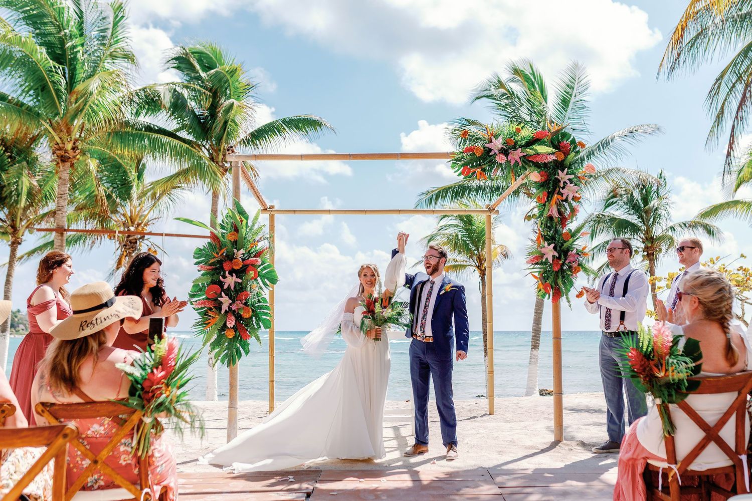 Nicole and Thomas celebrating beneath a tropical floral arch during their intimate Blue Venado beach wedding in Playa del Carmen.