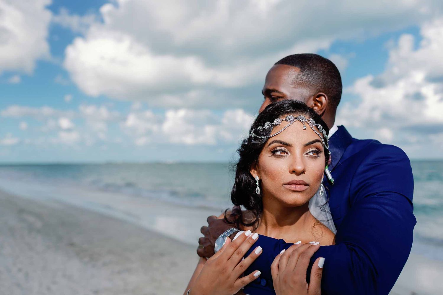 Elegant wedding couple portrait in Tulum on the beach with Caribbean ocean backdrop, bride wearing jeweled headpiece and groom embracing her