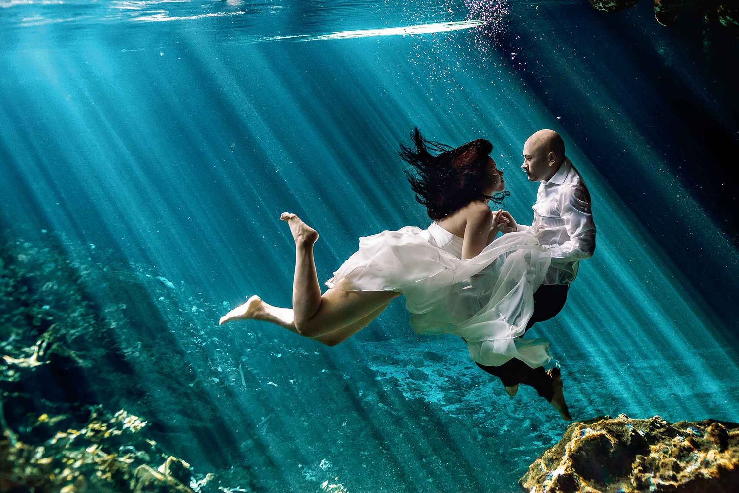 Bride and groom floating underwater in a Cancun cenote with dramatic sun rays during a destination wedding portrait session.
