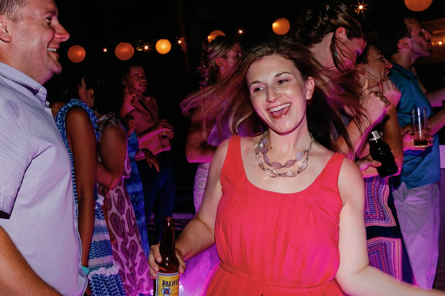 A guest in a coral dress dances with a bottle in her hand at the lantern-lit Tulum reception.