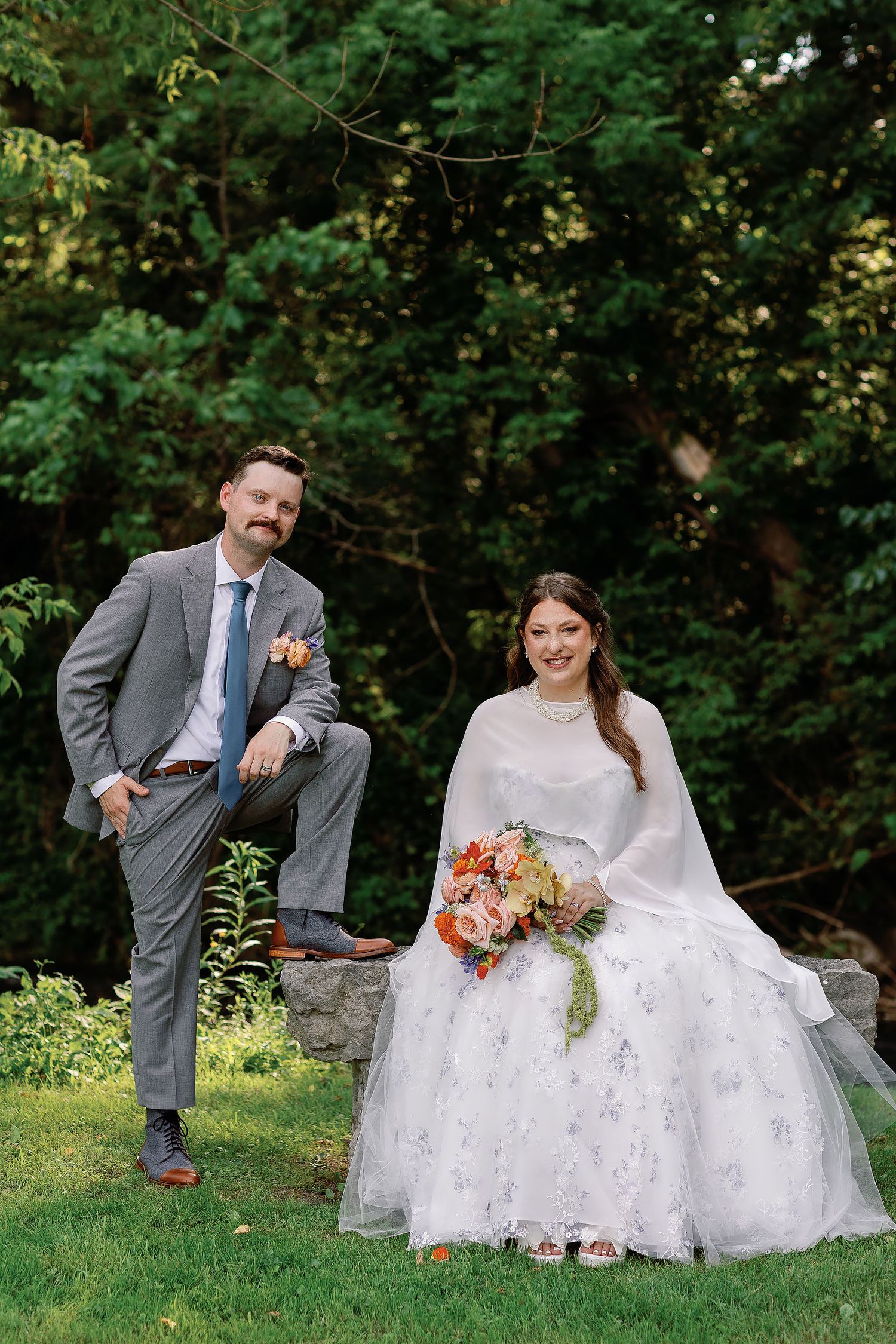 Sarah seated on a stone bench holding a colorful bouquet, with Stephen standing beside her during their Minnesota wedding portraits.