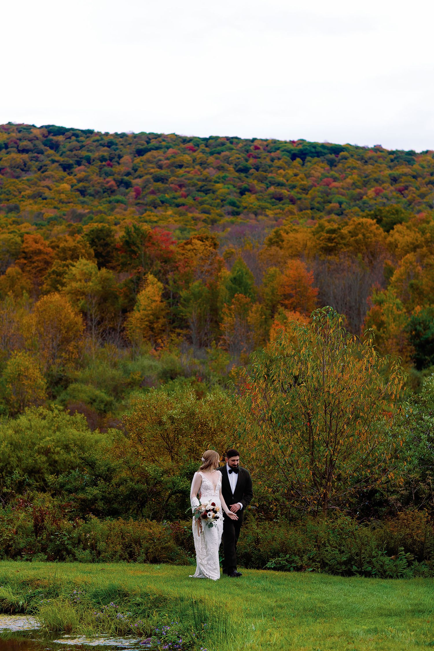 Natasha and Vladimir in wedding attire beside a pond, with a hillside of autumn foliage behind them in Duluth, Minnesota.