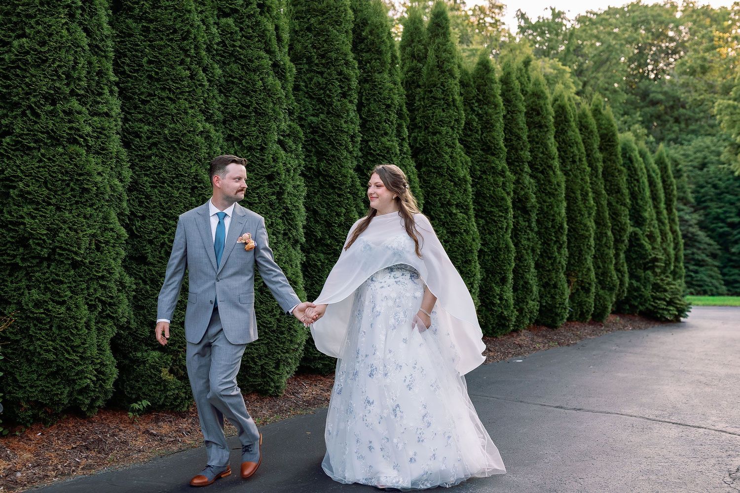 Sarah and Stephen holding hands and looking at each other as they walk beside tall evergreen trees in their wedding attire.