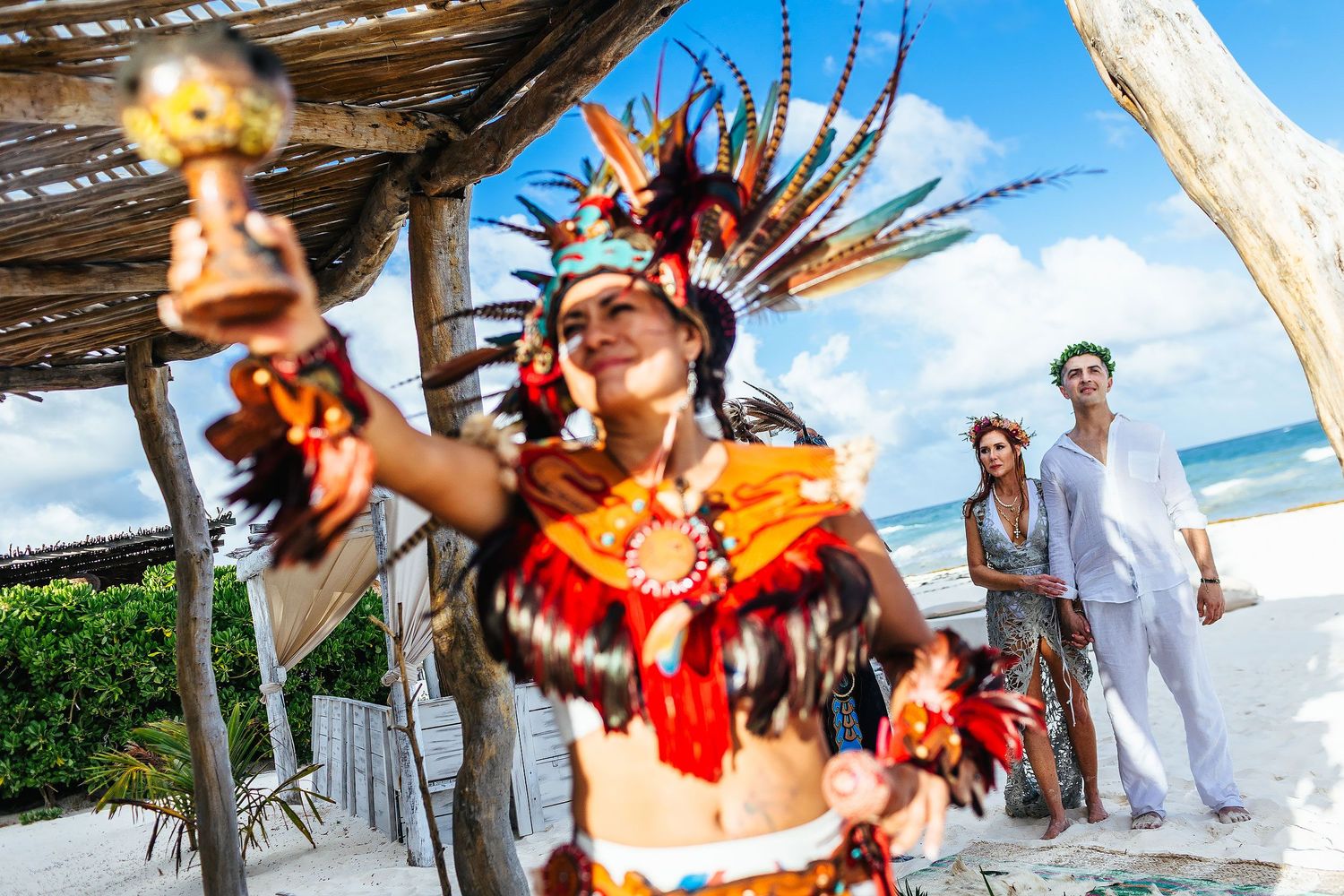 A ceremonial participant raises an instrument while Sarah and Evish watch from the beach.