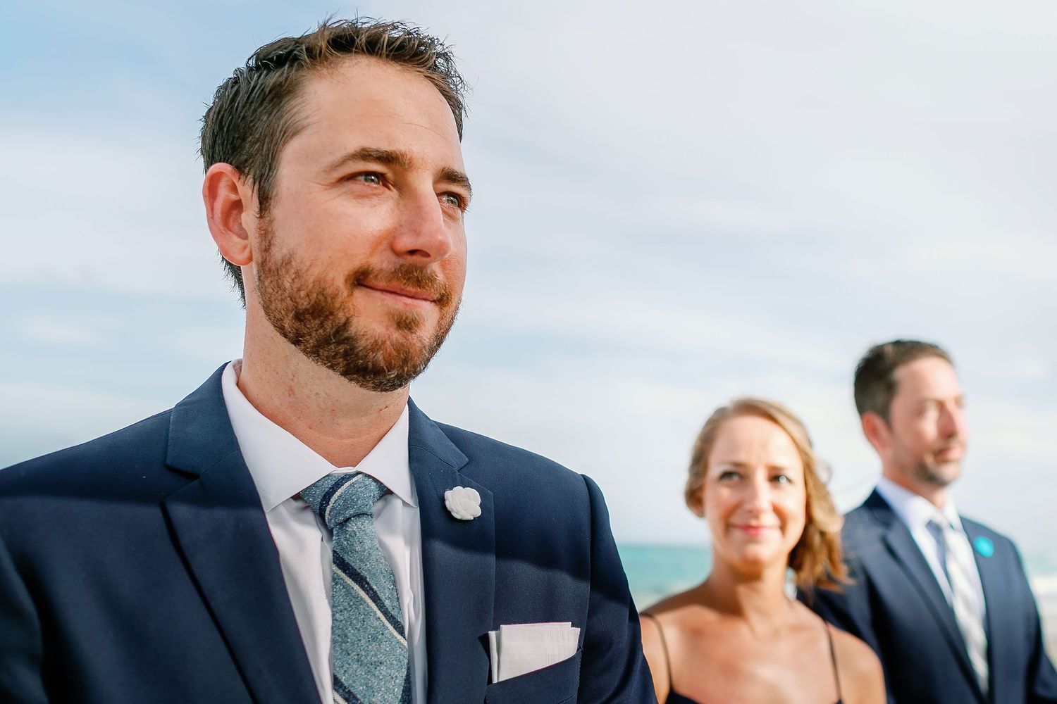 Matt waits for Ashley at the beachfront wedding ceremony with loved ones behind him.