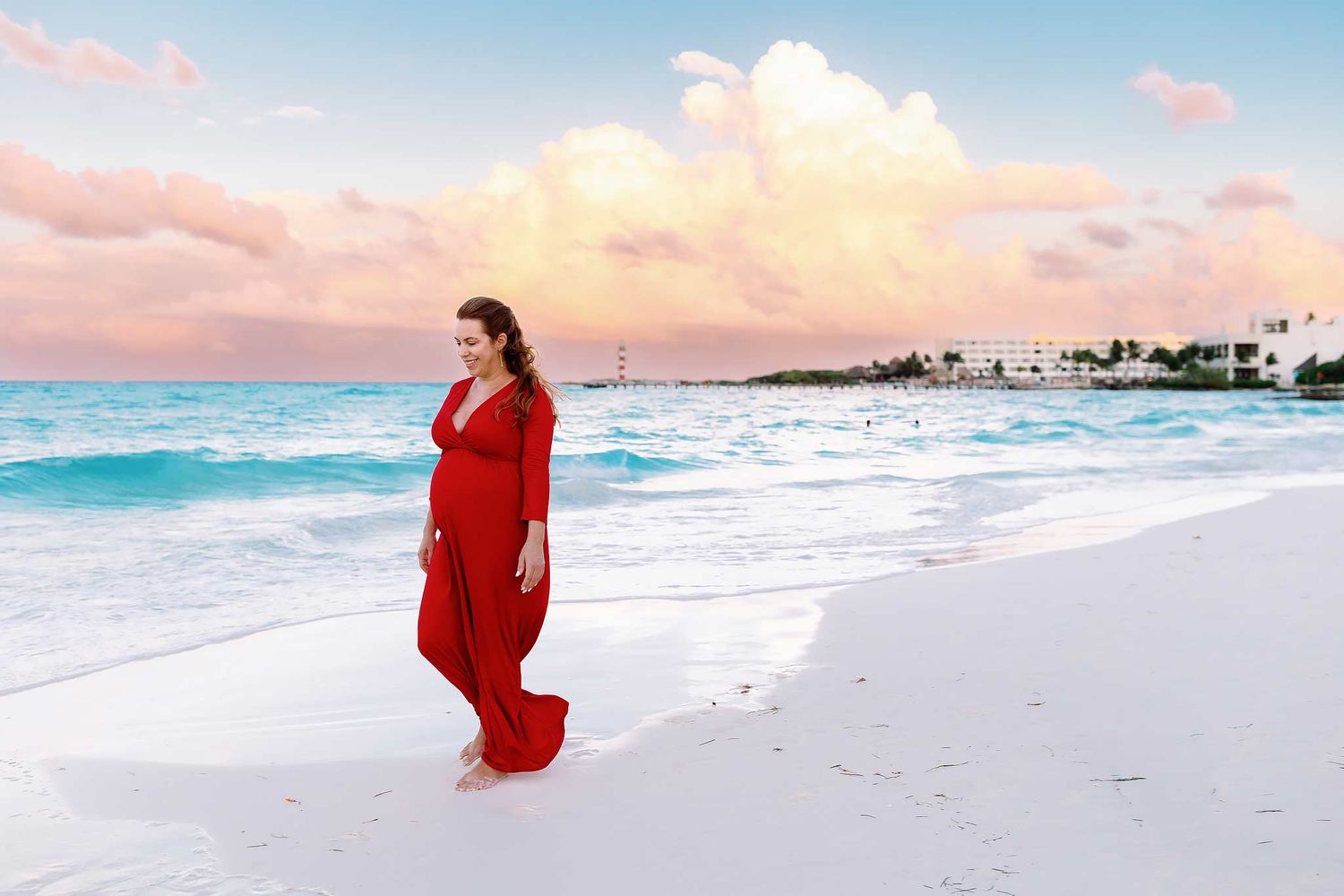 Pregnant woman in a flowing red dress walking along the white sand beach during a Cancun maternity photography session at sunset by the Cari
