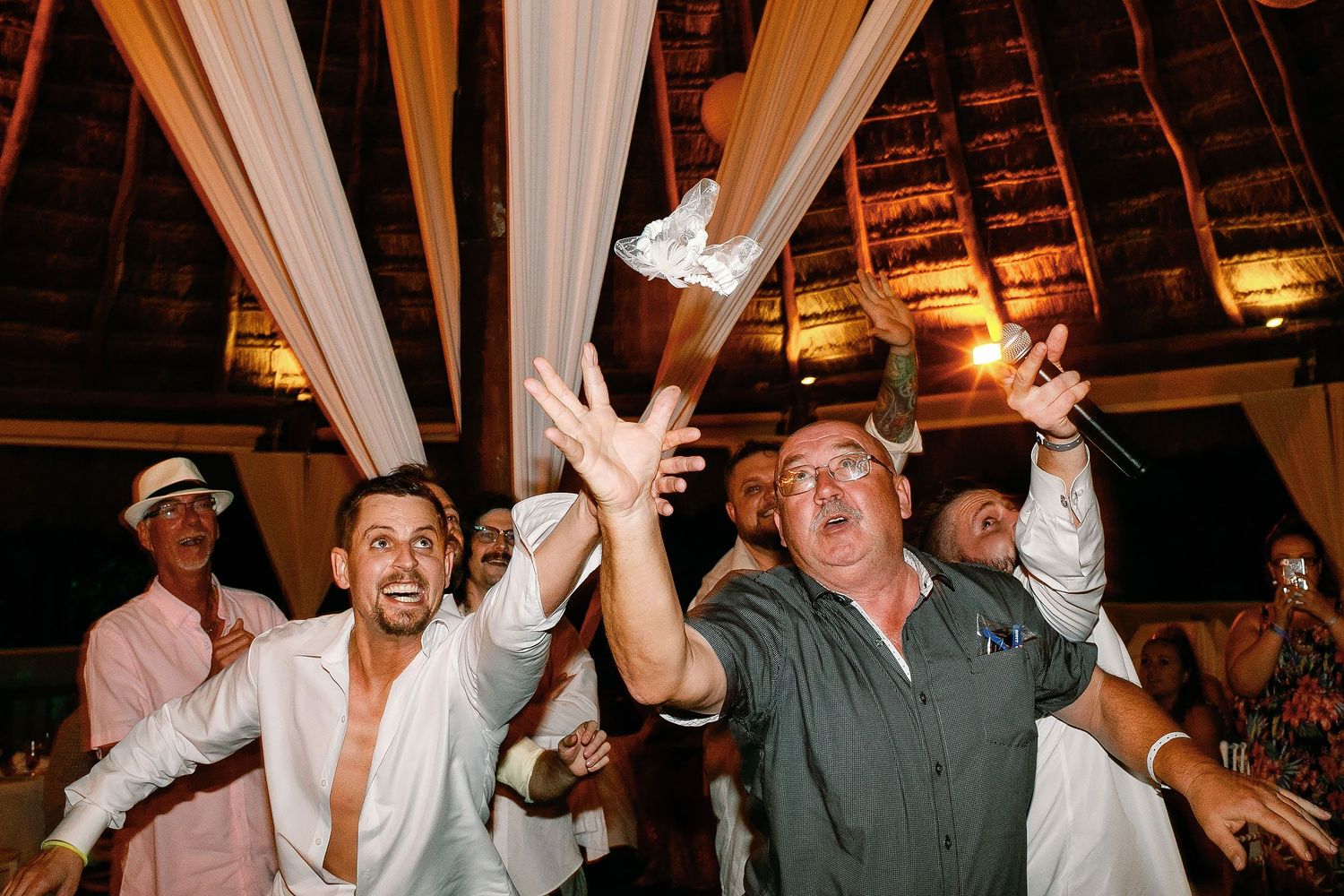 Wedding guests reaching to catch the garter inside the oceanfront palapa