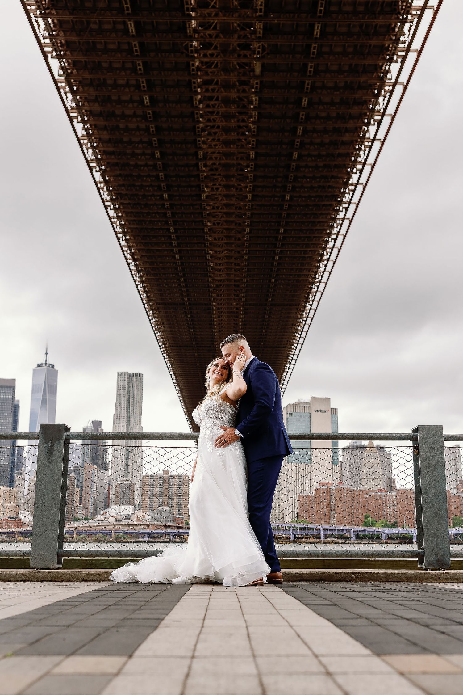 Klaudia and Matthew embracing in wedding attire beneath a bridge in Brooklyn, New York, with the Manhattan skyline behind them.