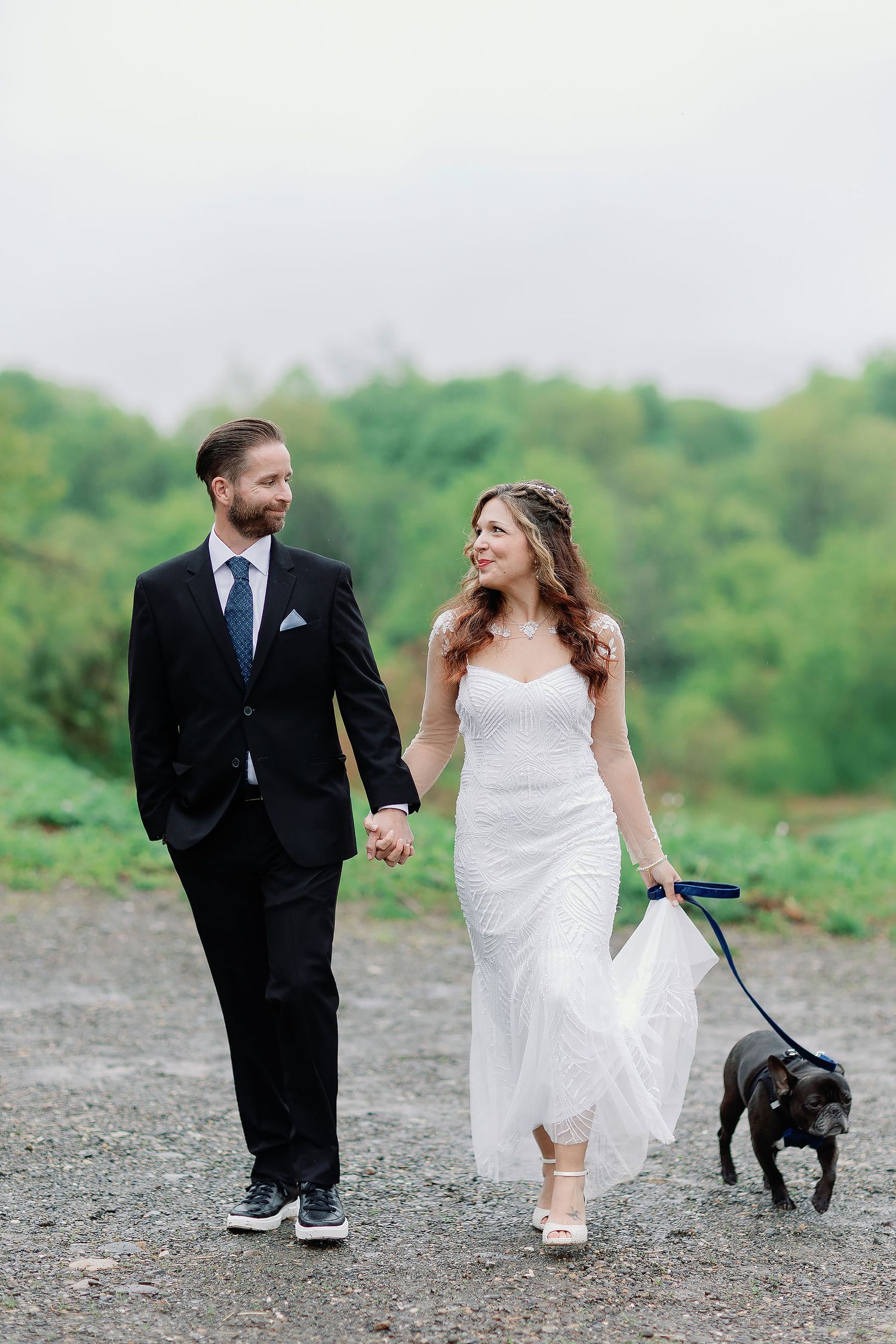 Bride and groom holding hands and walking their dog along a gravel path in Iowa, with green trees behind them.