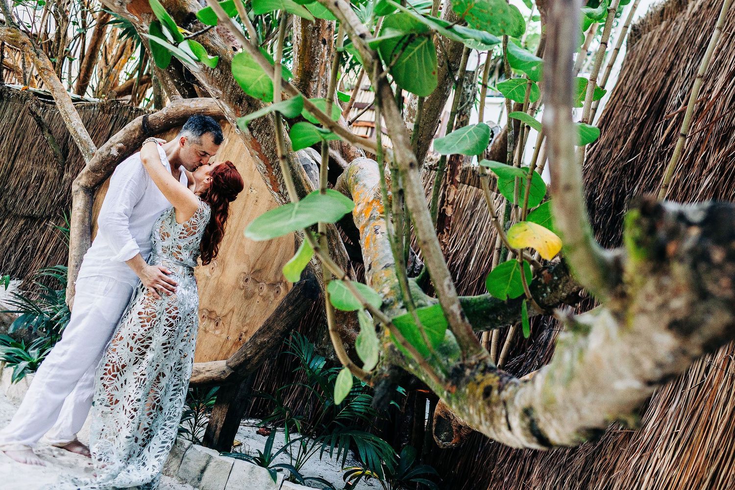 Sarah and Evish kiss among trees and rustic architecture in Tulum.