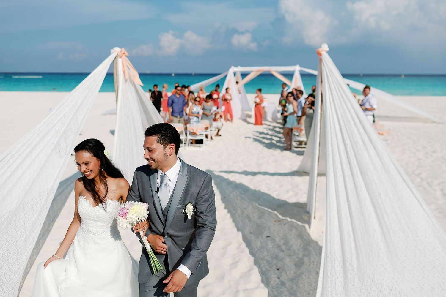 Felicitas and Keith smile while walking together along the sandy ceremony aisle.