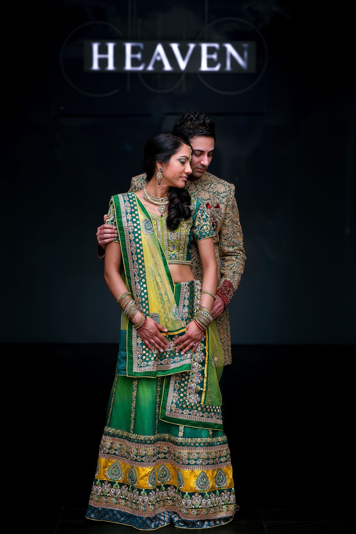 Bride and groom embracing at an Indian wedding in Minneapolis, wearing ornate green and gold wedding attire beneath an illuminated “HEAVEN”