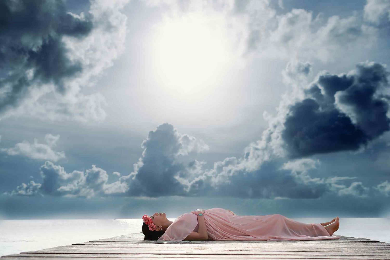 Pregnant woman in a flowing pink dress lying on a wooden dock over the Caribbean Sea during a dramatic Cancun maternity photography session.