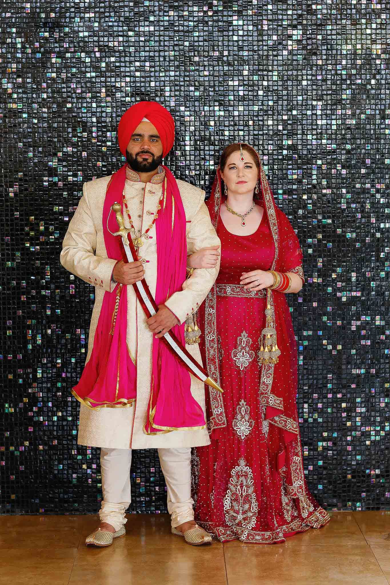 Traditional Sikh Indian wedding couple portrait with groom wearing turban and ceremonial sword beside bride in red lehenga during luxury des