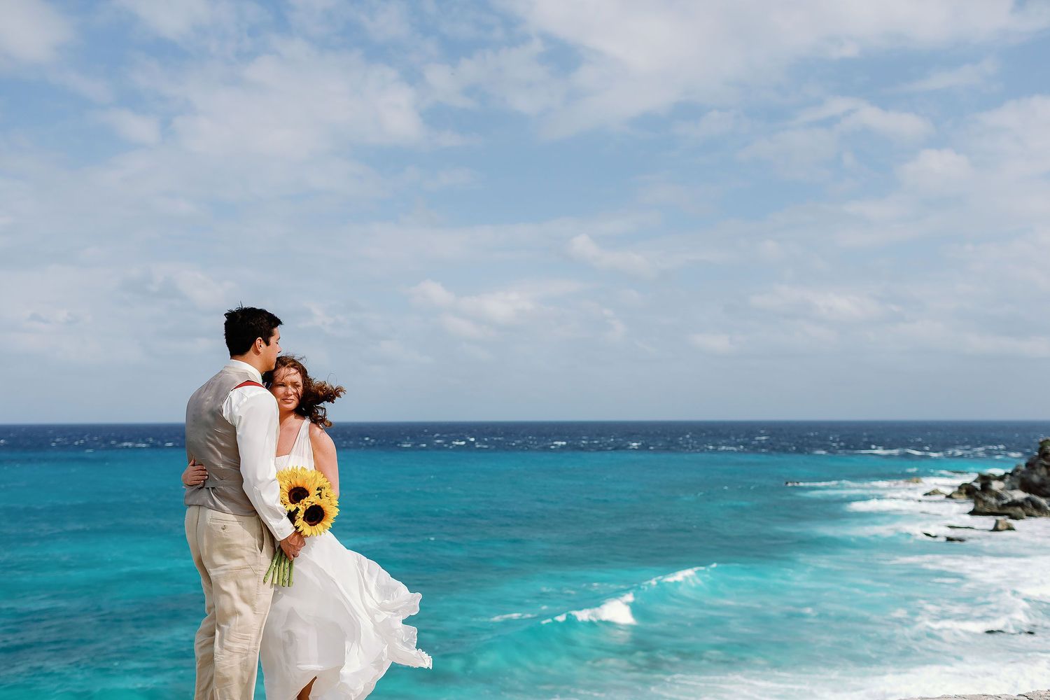 Li and Carlos pose above the turquoise Caribbean as the wind lifts her dress and sunflower bouquet.