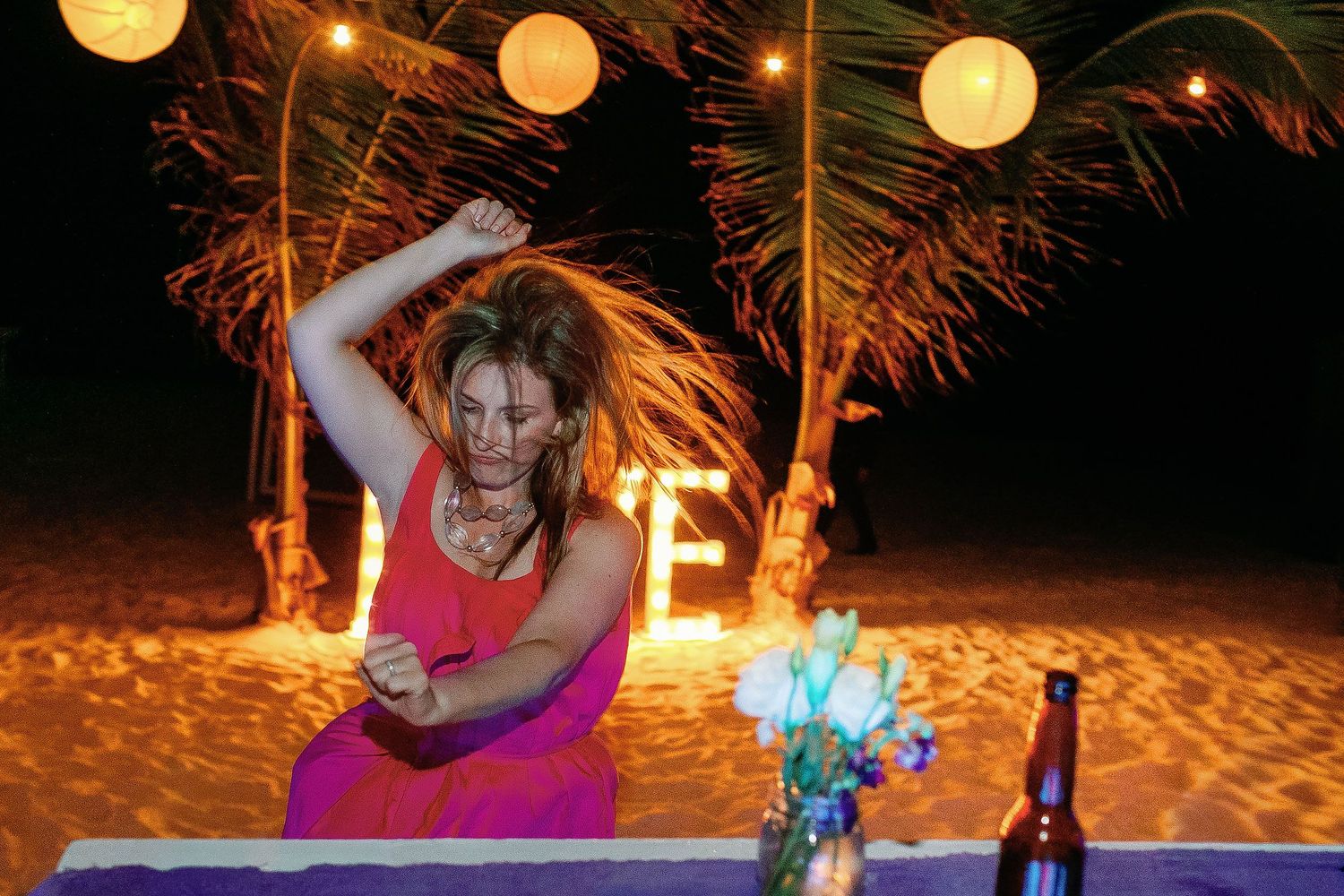 A guest in a bright coral dress dances beside the sand beneath glowing lanterns and palm trees.