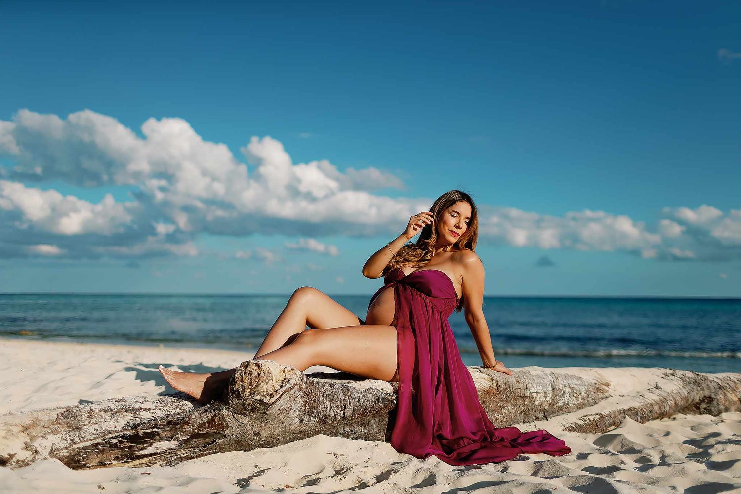 Pregnant woman in a flowing burgundy dress posing on driftwood along the beach during a Riviera Maya maternity photography session by the Ca