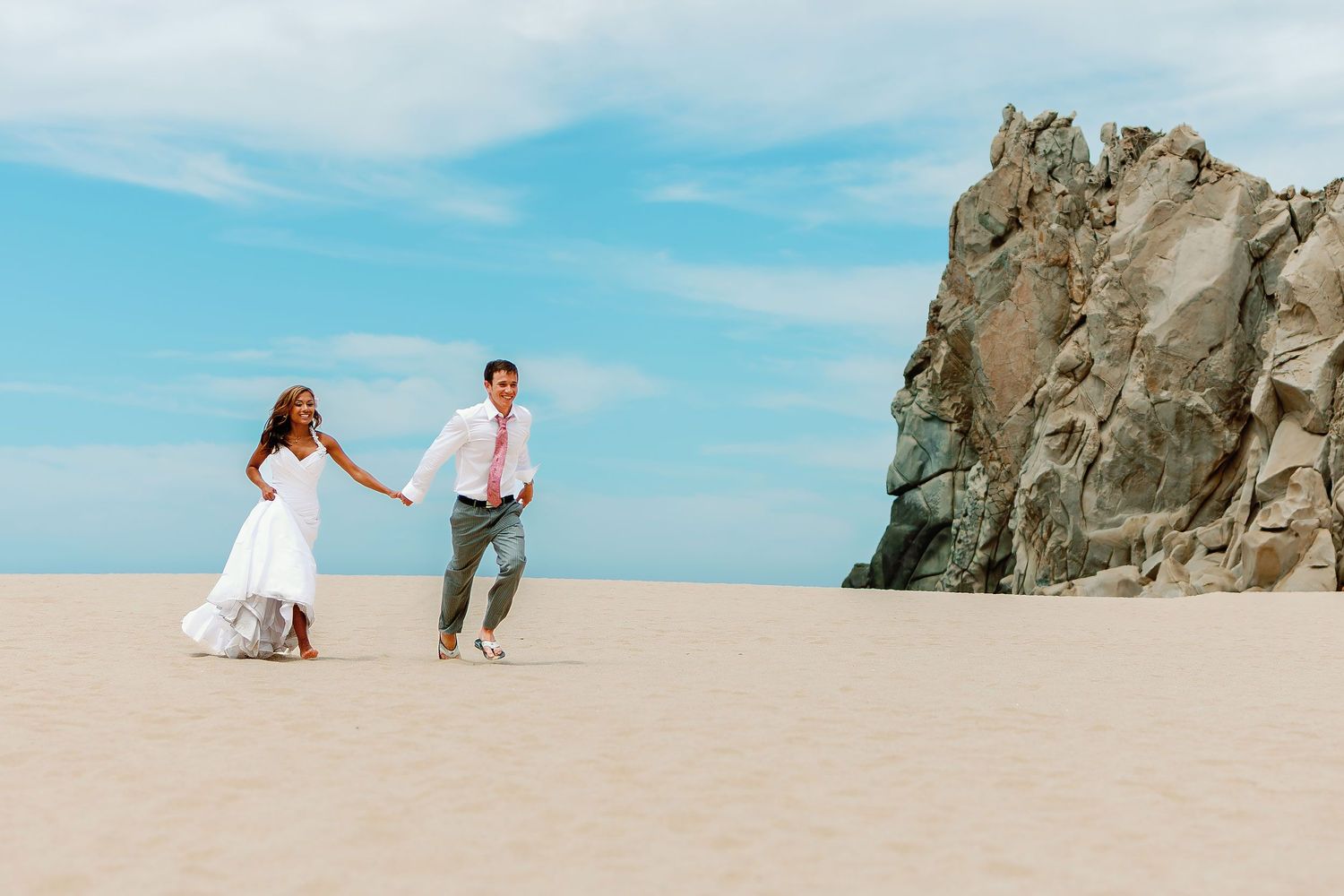 Bride and groom walking hand in hand on a secluded Los Cabos beach during a destination wedding portrait session.