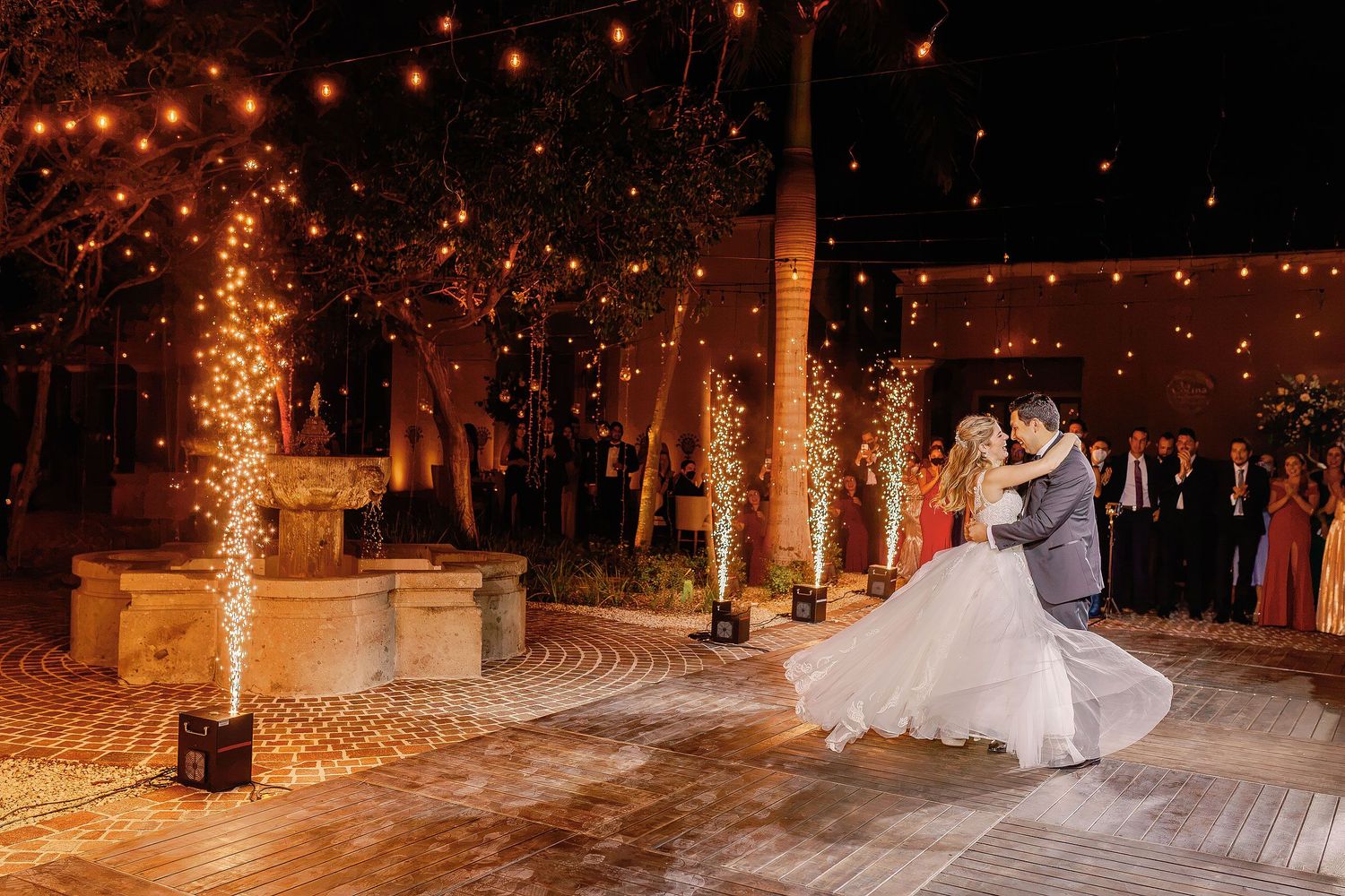Newlyweds sharing a first dance beneath string lights and sparkling fountains during their outdoor wedding reception at Rosewood Mayakoba