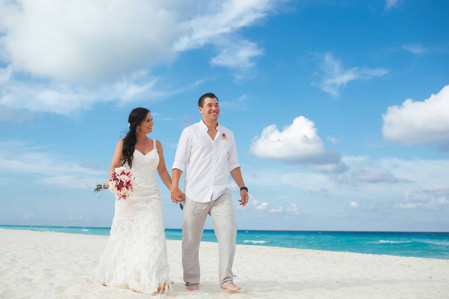 Bride and groom walking on a white sand beach in Cancun during a destination wedding with turquoise Caribbean water and blue sky.