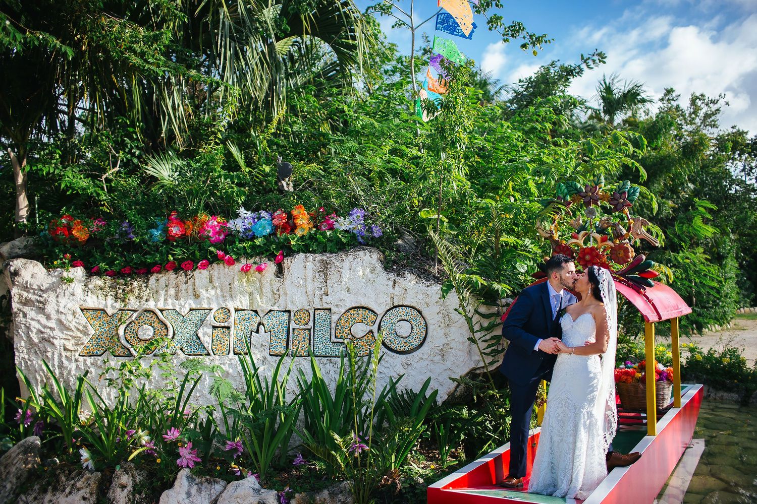 Bride and groom kissing beside the colorful Xoximilco sign and traditional trajinera during their Riviera Maya wedding portrait session.
