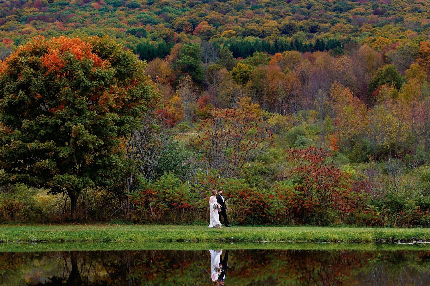 Natasha and Vladimir standing beside a pond in Duluth, Minnesota, with their wedding attire and the colorful autumn trees reflected in the w