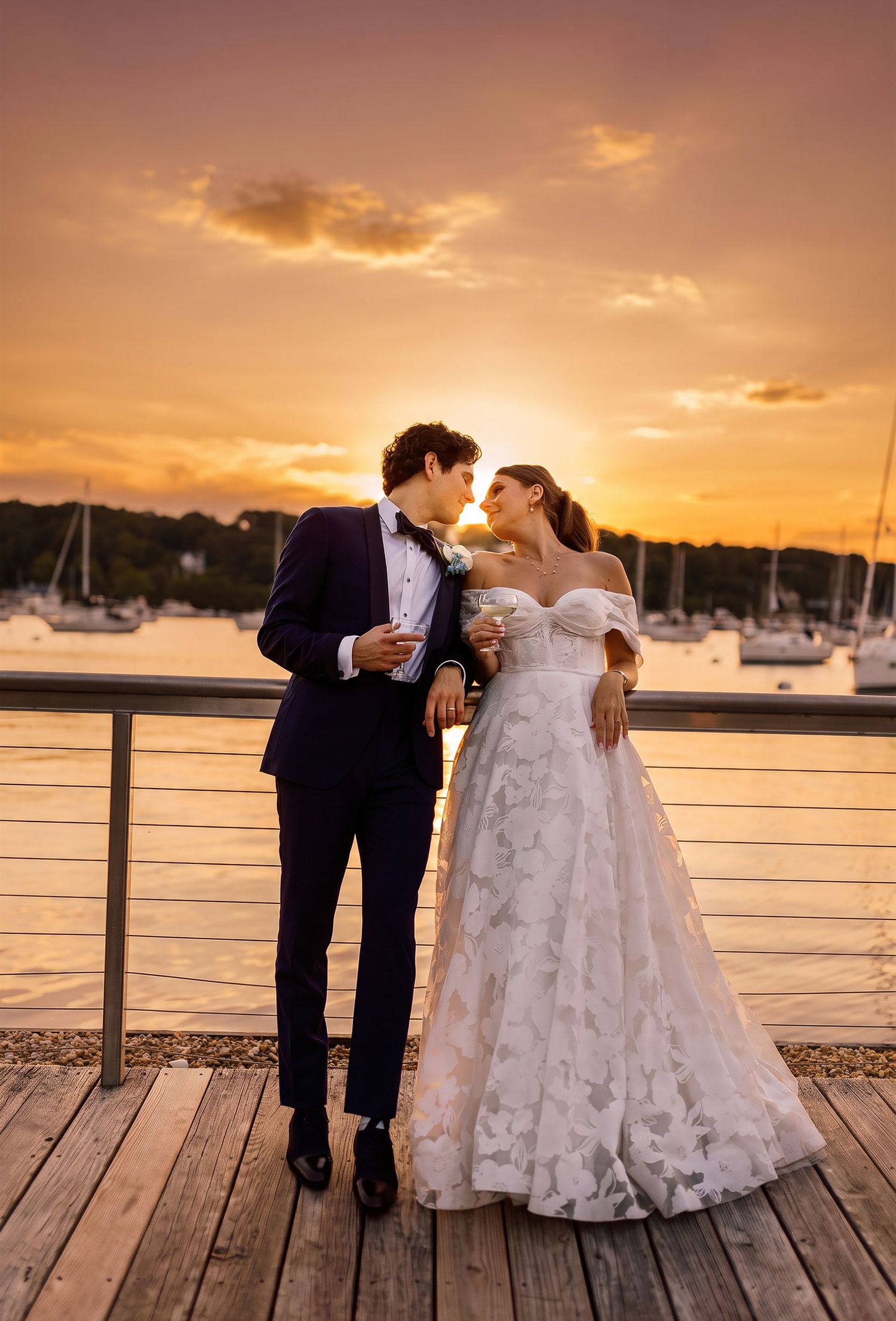 Bride and groom holding drinks beside a waterfront railing at their Illinois wedding, with sailboats and a golden sunset behind them.