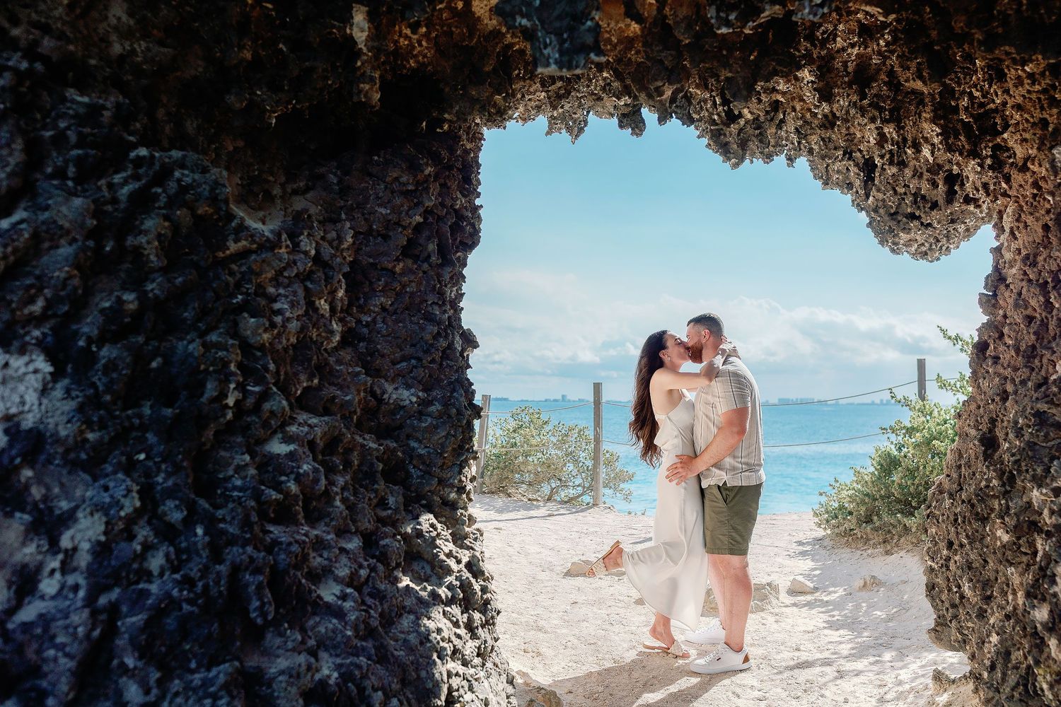 Alexia and Sam kiss beneath a natural rock arch with the Caribbean Sea behind them.